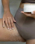 Close-up of woman using Bermuda Dark Spot Exfoliating Scrub on her thigh, salt-like exfoliant texture visible, skin care for dark spots